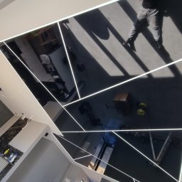 Sławomir Owerko - Błyszczący, czarny sufit z wbudowanymi liniami LED tworzącymi geometryczny wzór, odbijający wnętrze pomieszczenia z widoczną osobą i narzędziami.