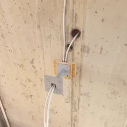 Szare kable elektryczne prowadzone wewnątrz białych rurek, wychodzące ze skrzynki montażowej z metalowym mocowaniem do sufitu z surowego betonu.