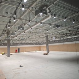 MD-Elektro Maciej Doniec - Rozległa hala z równomiernie rozmieszczonymi, cylindrycznymi lampami sufitowymi i systemem wentylacji. Widoczne koryta kablowe i instalacje pod stropem.