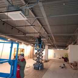 MD-Elektro Maciej Doniec - Montaż instalacji elektrycznej w nowym lokalu: pracownicy na podnośnikach instalują koryta kablowe i oświetlenie pod sufitem. Widoczne kable i narzędzia.