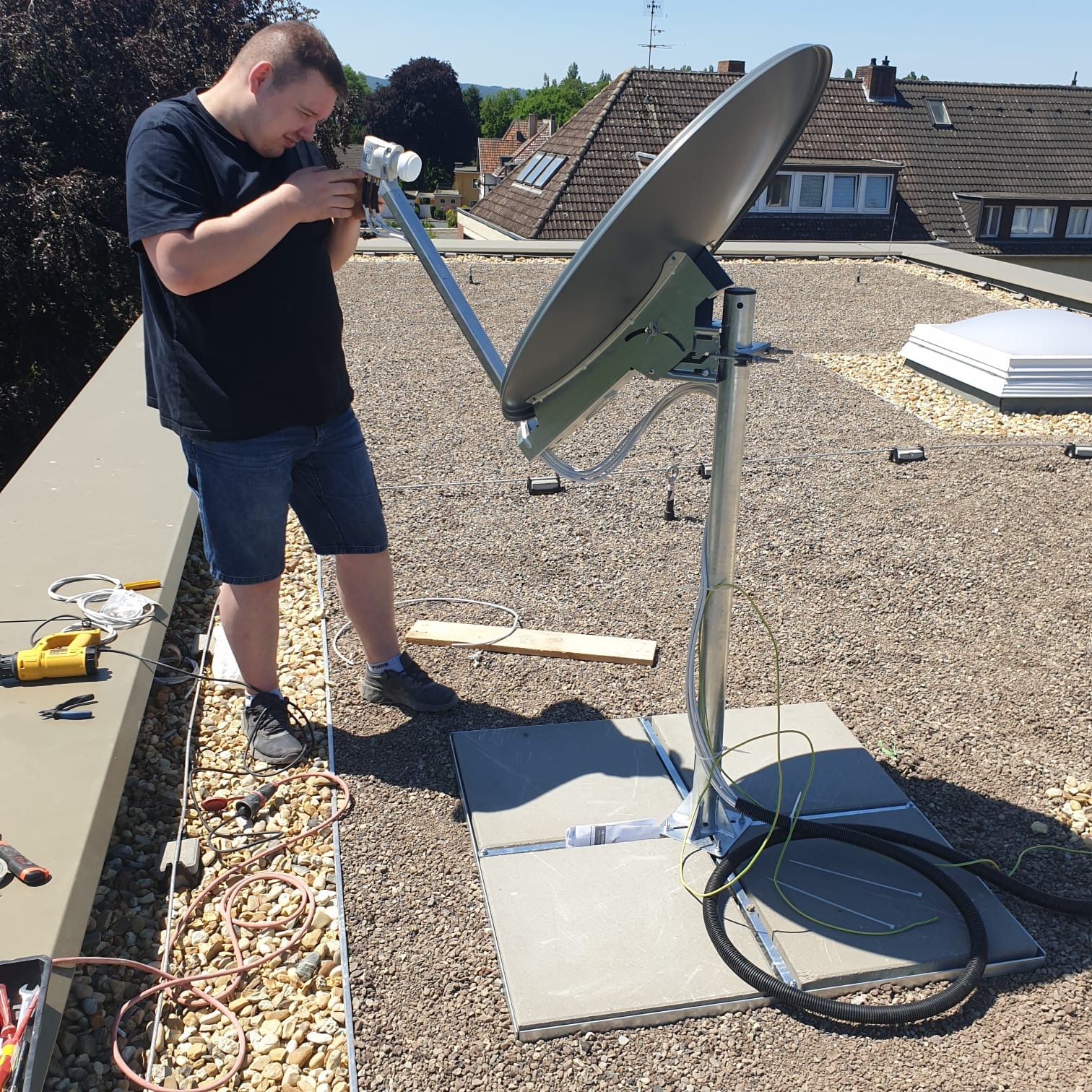 Instalator w trakcie montażu anteny satelitarnej na płaskim dachu pokrytym żwirem, widoczne narzędzia i przewody.