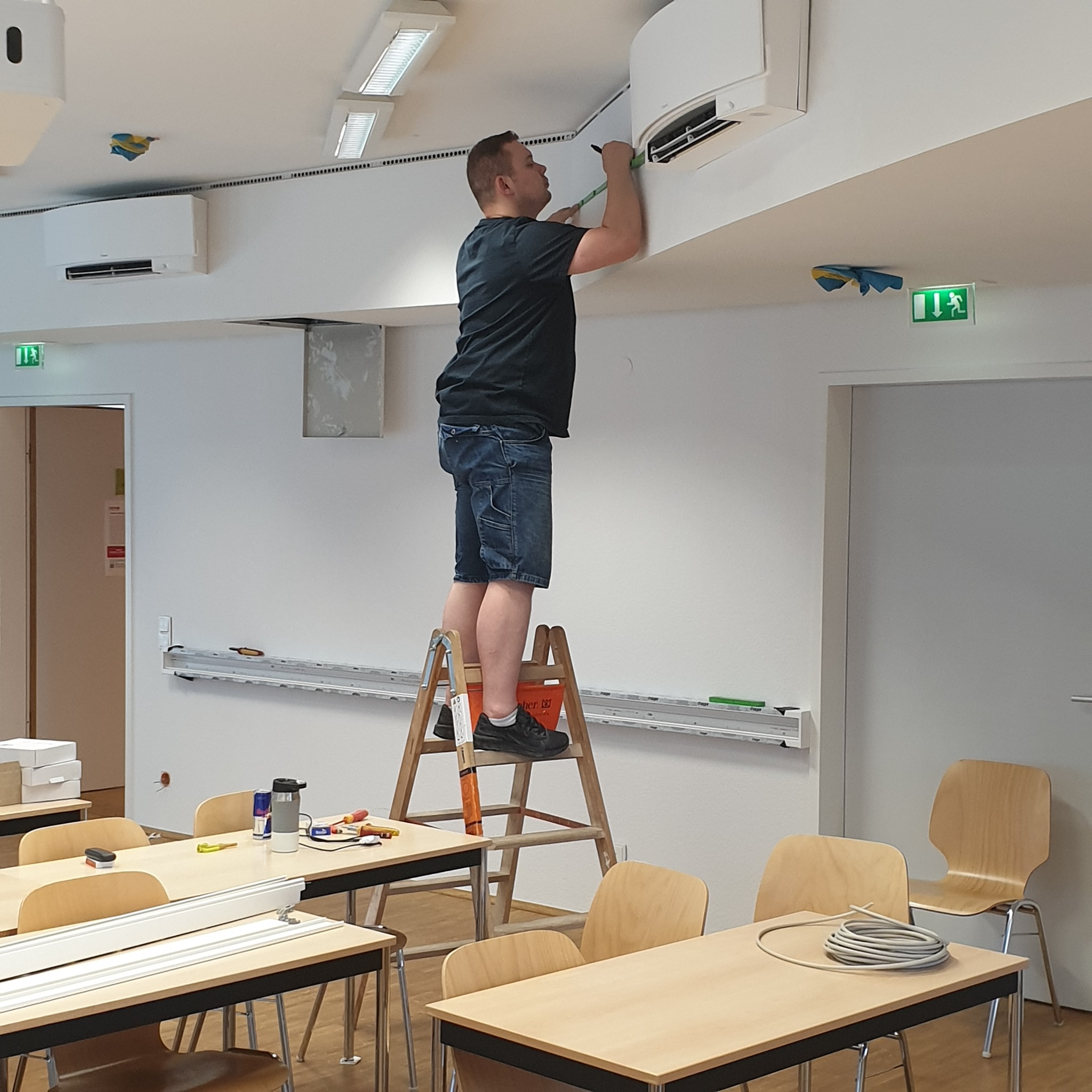 Mężczyzna na drabinie mierzy klimatyzator w jasnym pomieszczeniu z biurkami i krzesłami, widoczne narzędzia i przewody.