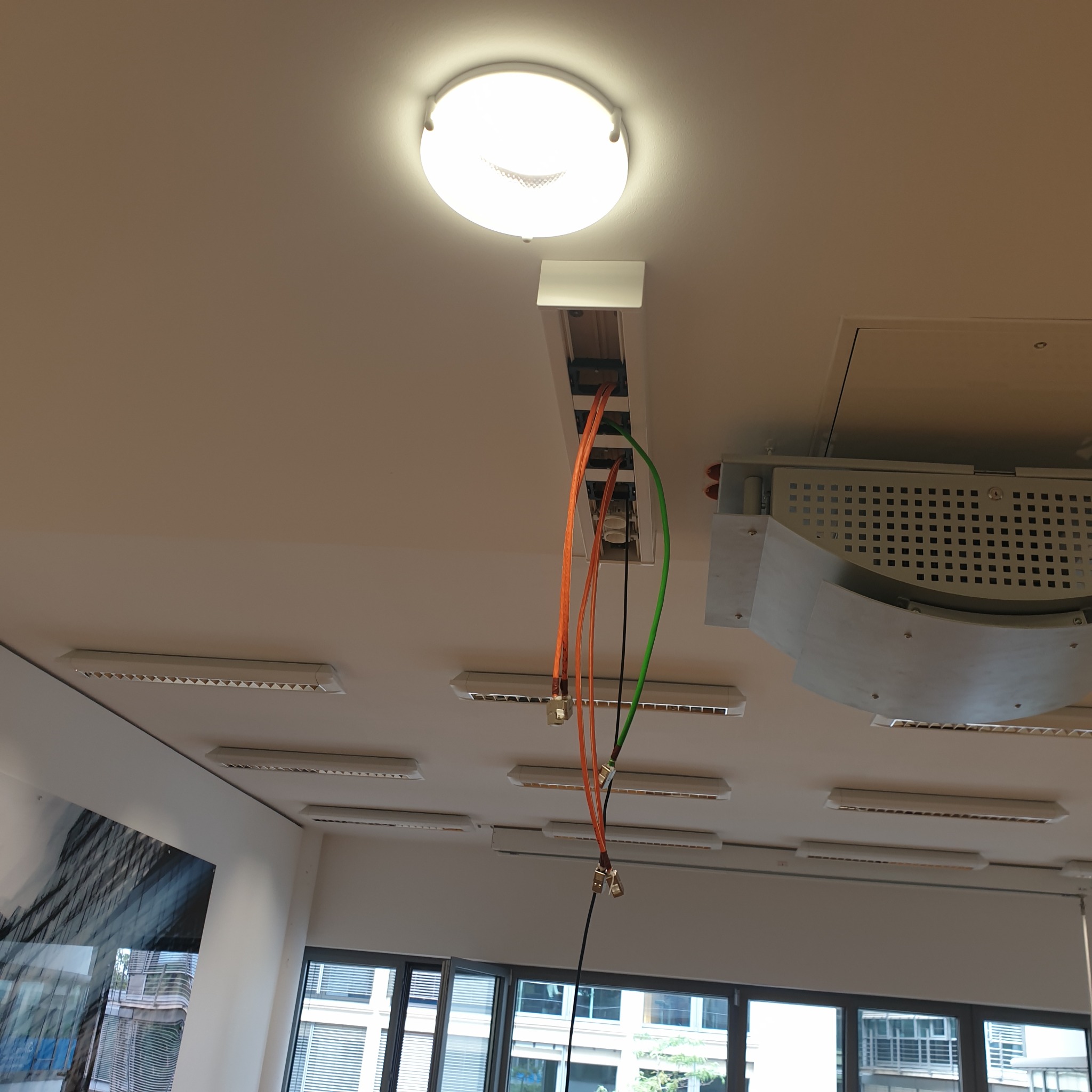 Widok na sufit z zamontowanym oświetleniem panelowym LED i otwartą listwą z przewodami elektrycznymi w kolorach pomarańczowym i zielonym, z widocznymi złączkami.