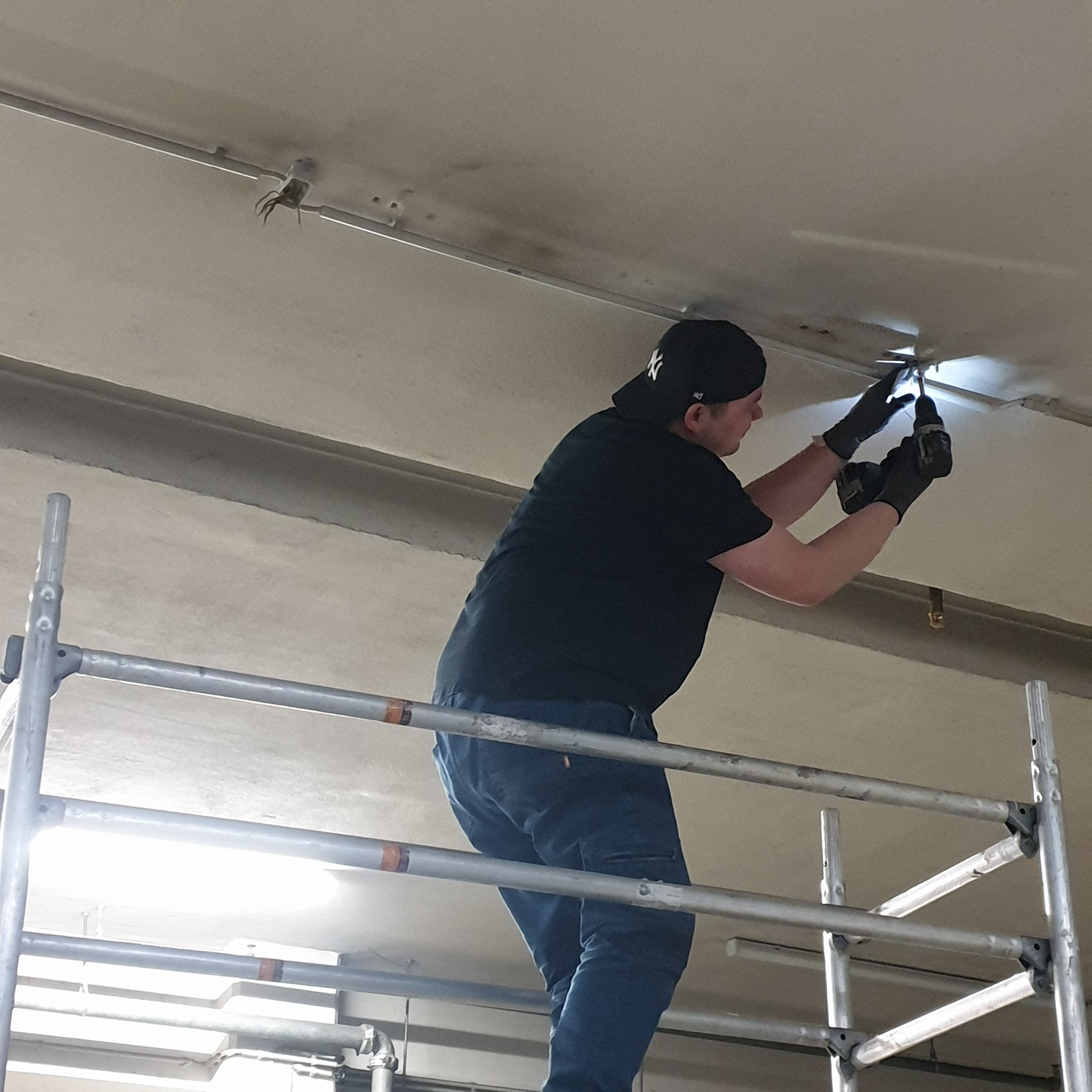 Elektryk na rusztowaniu w czarnych rękawicach i czapce z daszkiem instaluje przewody w suficie za pomocą wiertarki akumulatorowej.