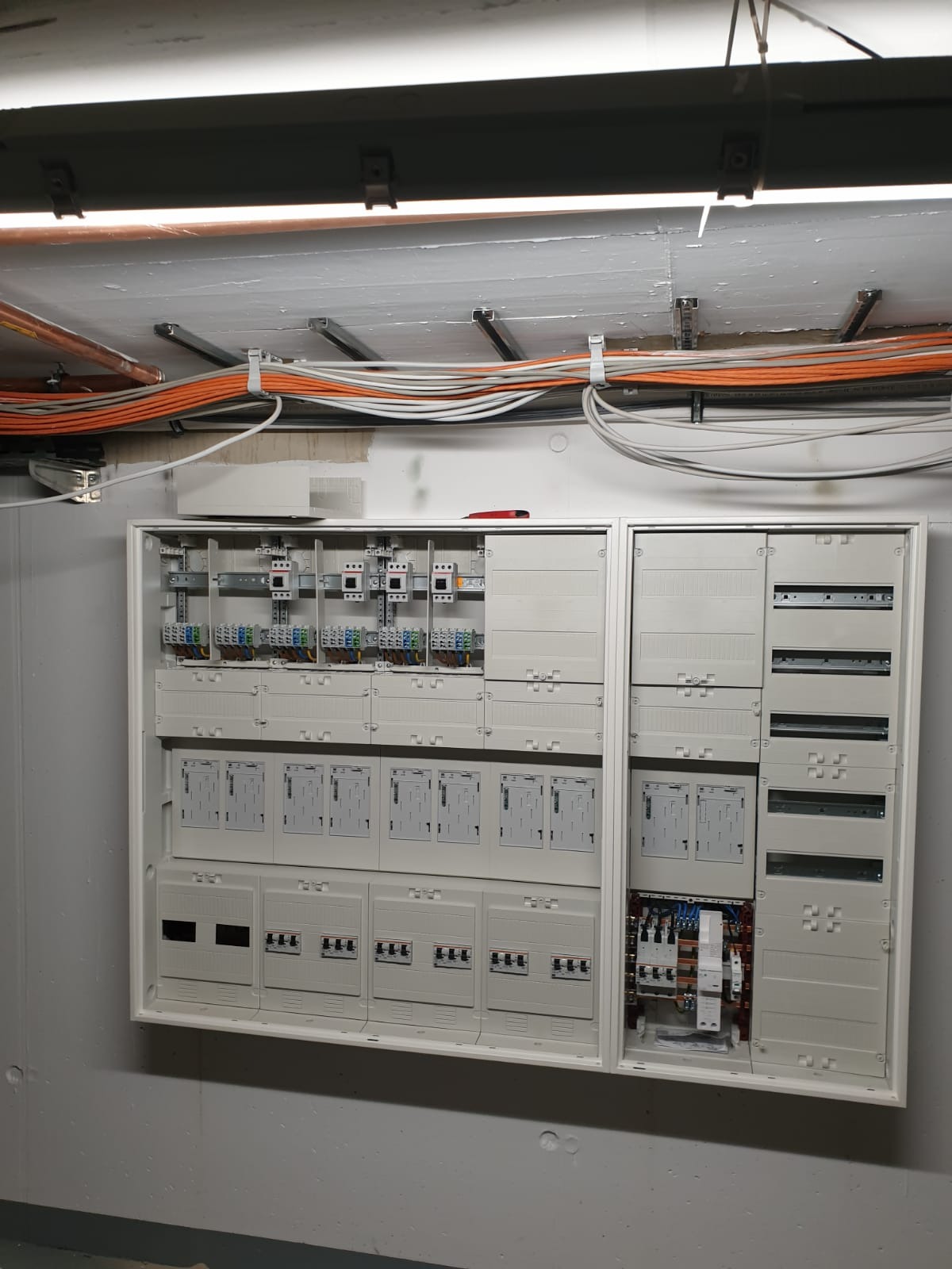 Rozdzielnica elektryczna zamontowana na ścianie, z widocznymi przewodami w listwach na suficie. Część obudowy rozdzielnicy jest otwarta, ukazując wewnętrzne okablowanie.