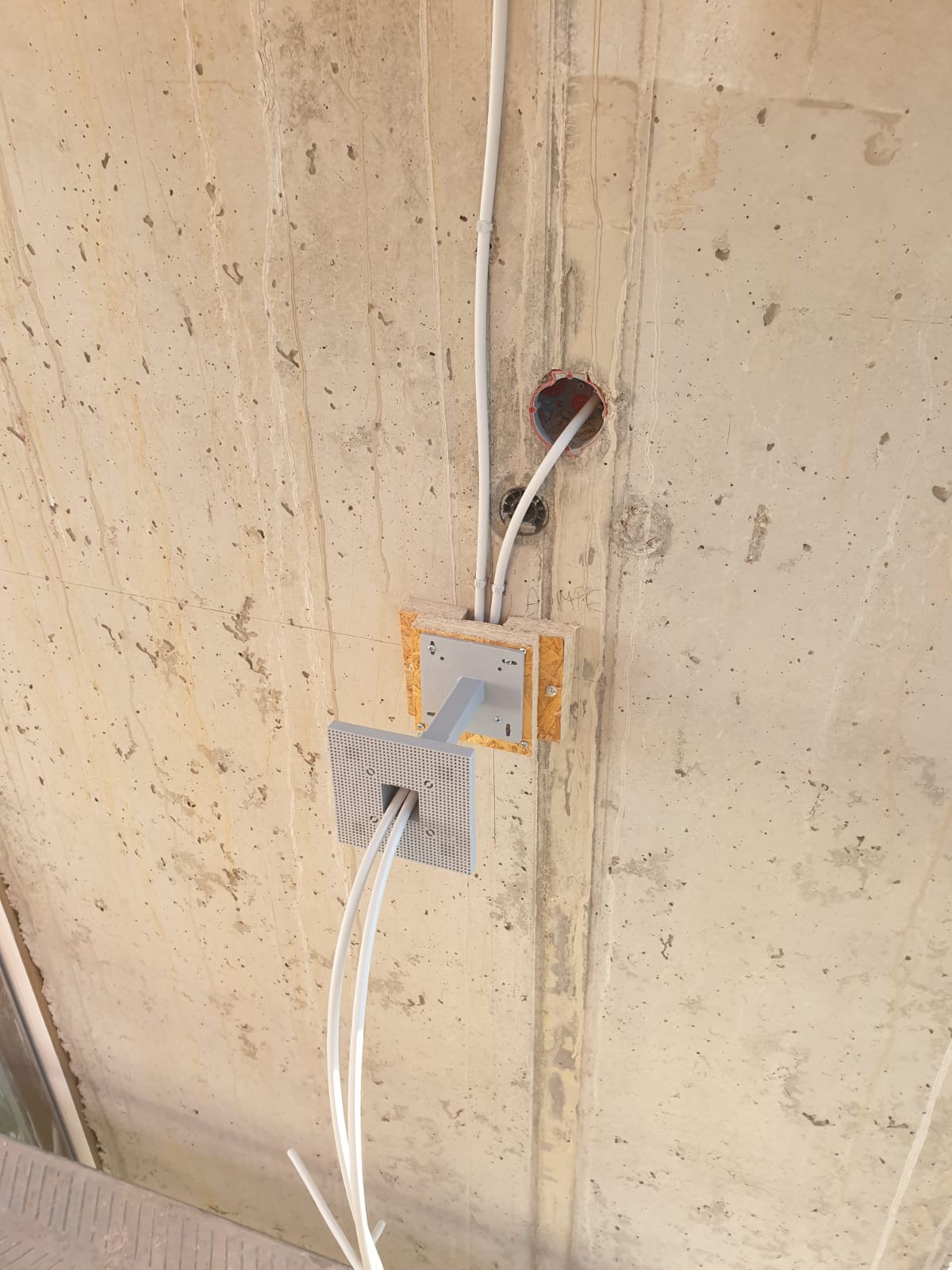 Szare kable elektryczne prowadzone wewnątrz białych rurek, wychodzące ze skrzynki montażowej z metalowym mocowaniem do sufitu z surowego betonu.