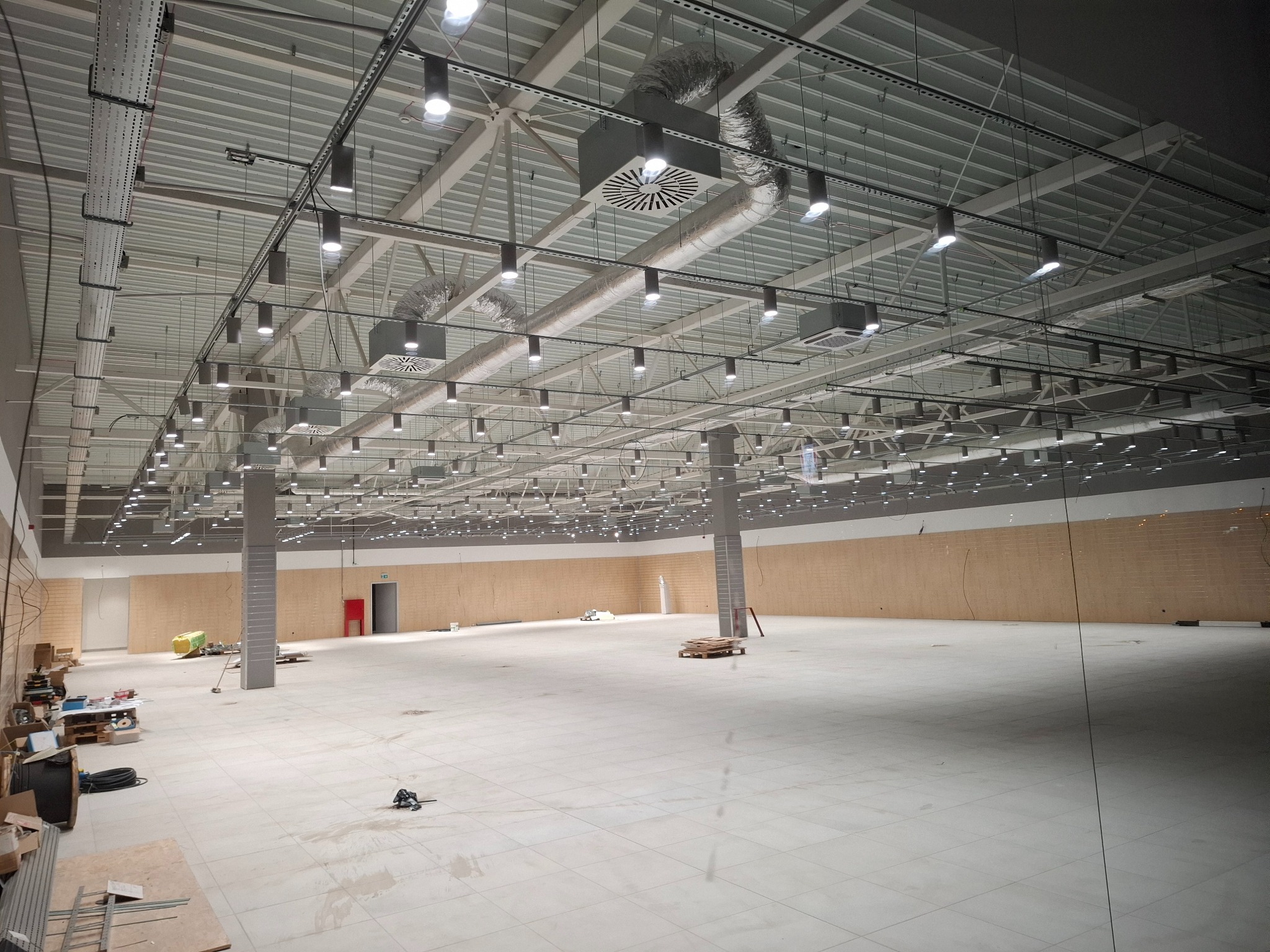 Rozległa hala z równomiernie rozmieszczonymi, cylindrycznymi lampami sufitowymi i systemem wentylacji. Widoczne koryta kablowe i instalacje pod stropem.