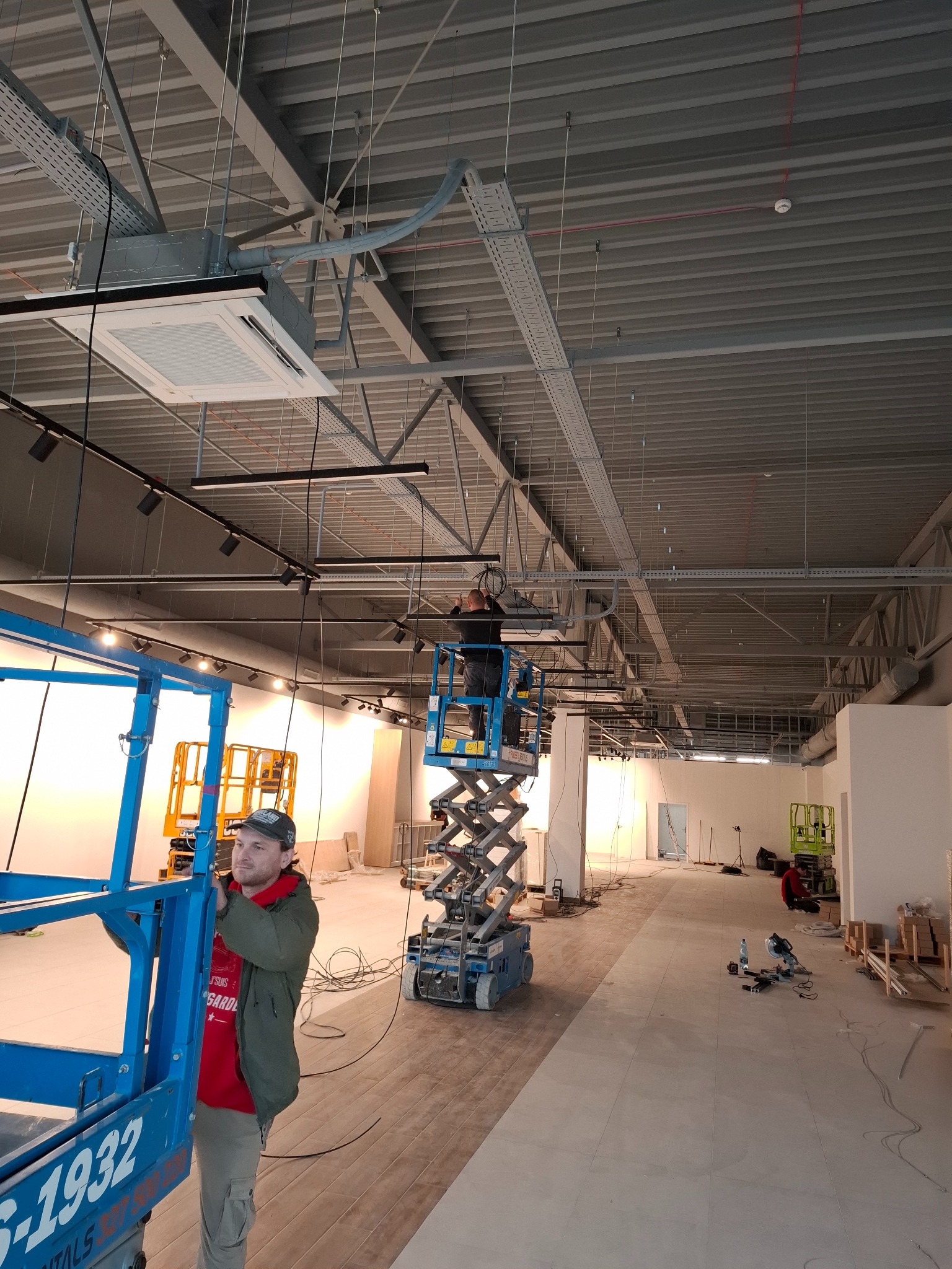 Montaż instalacji elektrycznej w nowym lokalu: pracownicy na podnośnikach instalują koryta kablowe i oświetlenie pod sufitem. Widoczne kable i narzędzia.