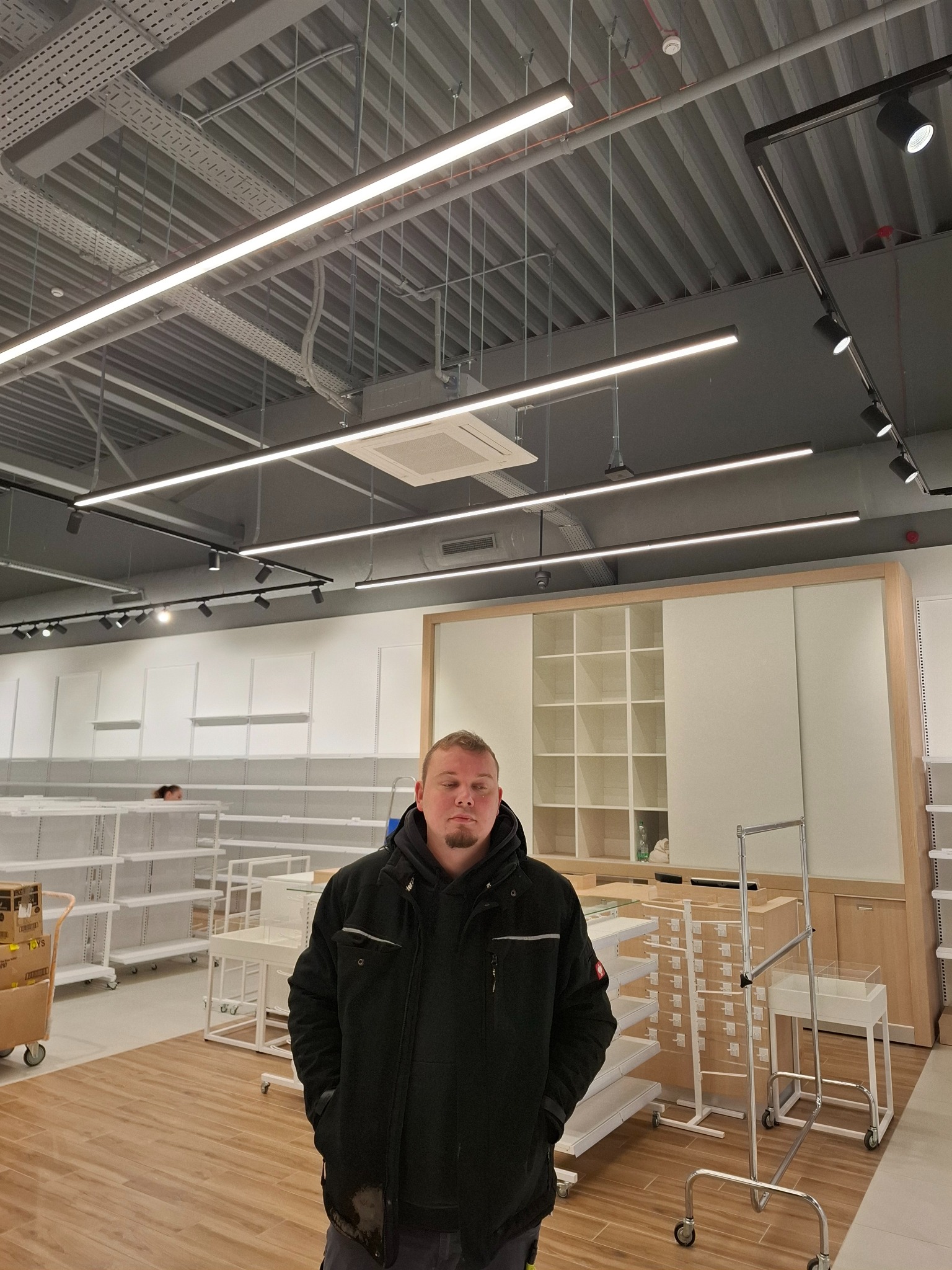 Nowoczesne oświetlenie LED w nowym sklepie, widoczne koryta kablowe pod sufitem. Mężczyzna w kurtce na pierwszym planie. Jasne wnętrze z regałami.