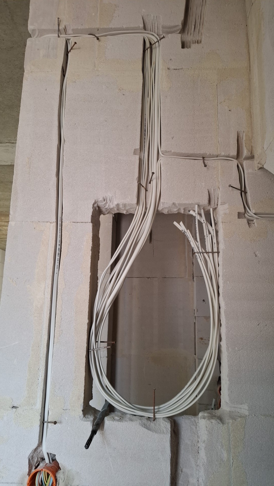 Białe kable elektryczne ułożone w bruzdach w ścianie z białego kamienia, częściowo zabezpieczone gwoździami. Na dole widoczna pomarańczowa puszka instalacyjna.