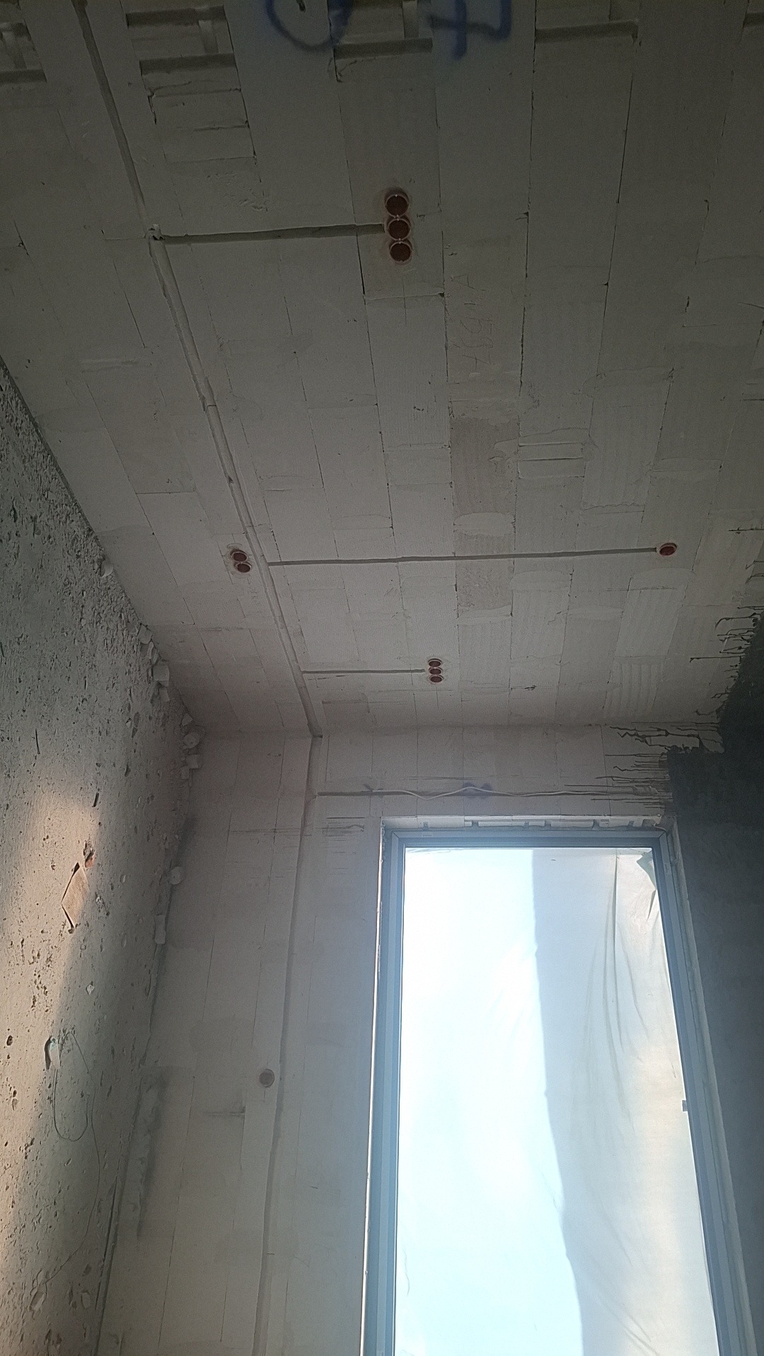 Surowy strop z widocznymi przewodami elektrycznymi i puszkami instalacyjnymi. Widok z dołu, okno w tle.