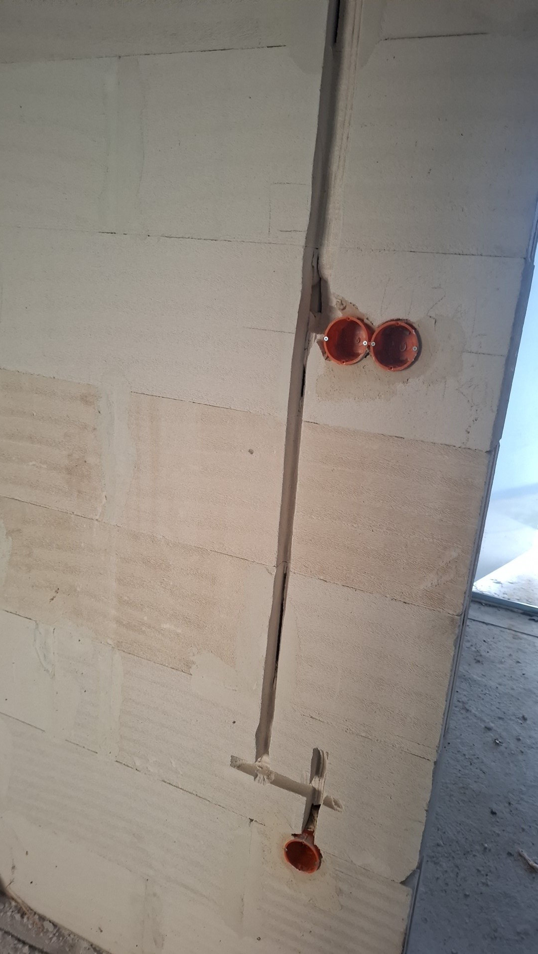 Biała ściana z wyżłobionym pionowym rowkiem, w którym umieszczono pomarańczowe puszki elektryczne, prezentujące etap prac instalacji elektrycznej.