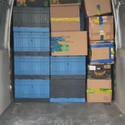Wnętrze busa wypełnione kartonami i niebieskimi skrzynkami, gotowe do transportu. Widok z tyłu pojazdu, podłoga lekko zabrudzona.