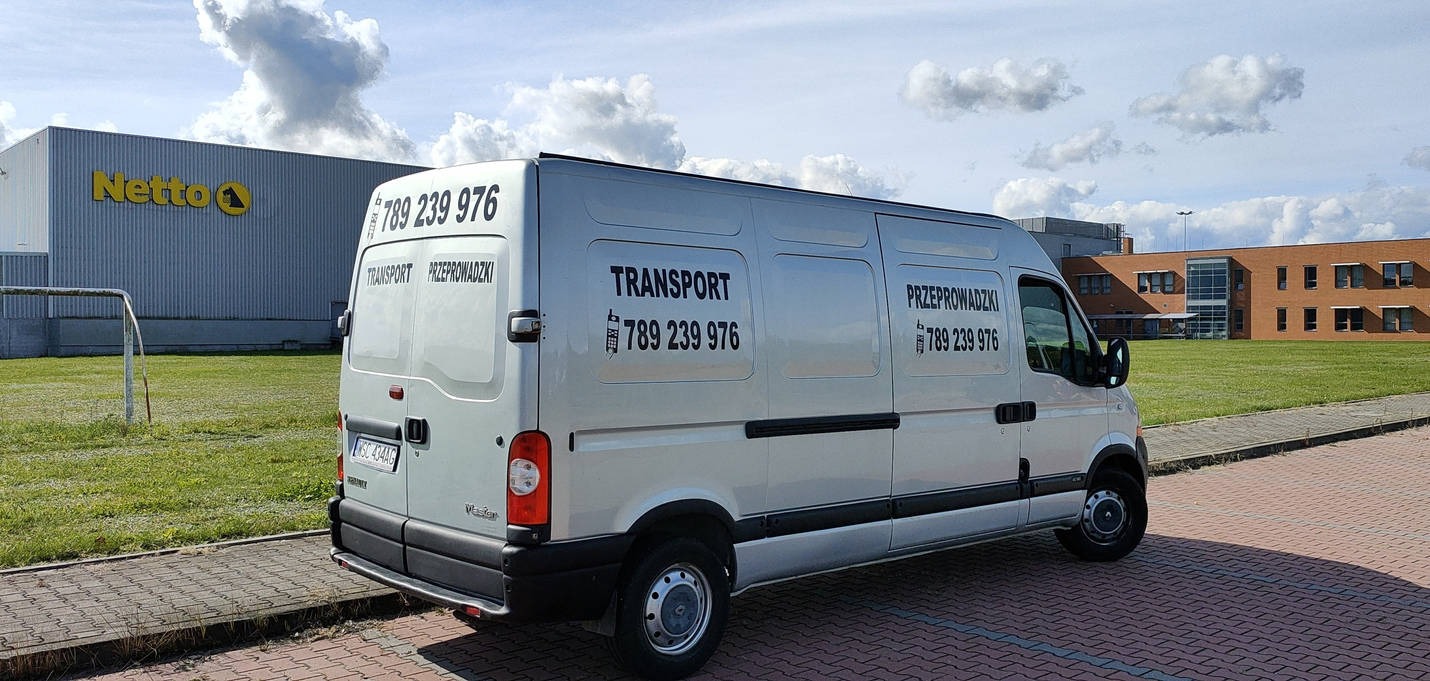 Srebrny bus z napisem 'TRANSPORT PRZEPROWADZKI' i numerem telefonu 789 239 976, zaparkowany na tle budynku Netto i budynku biurowego.