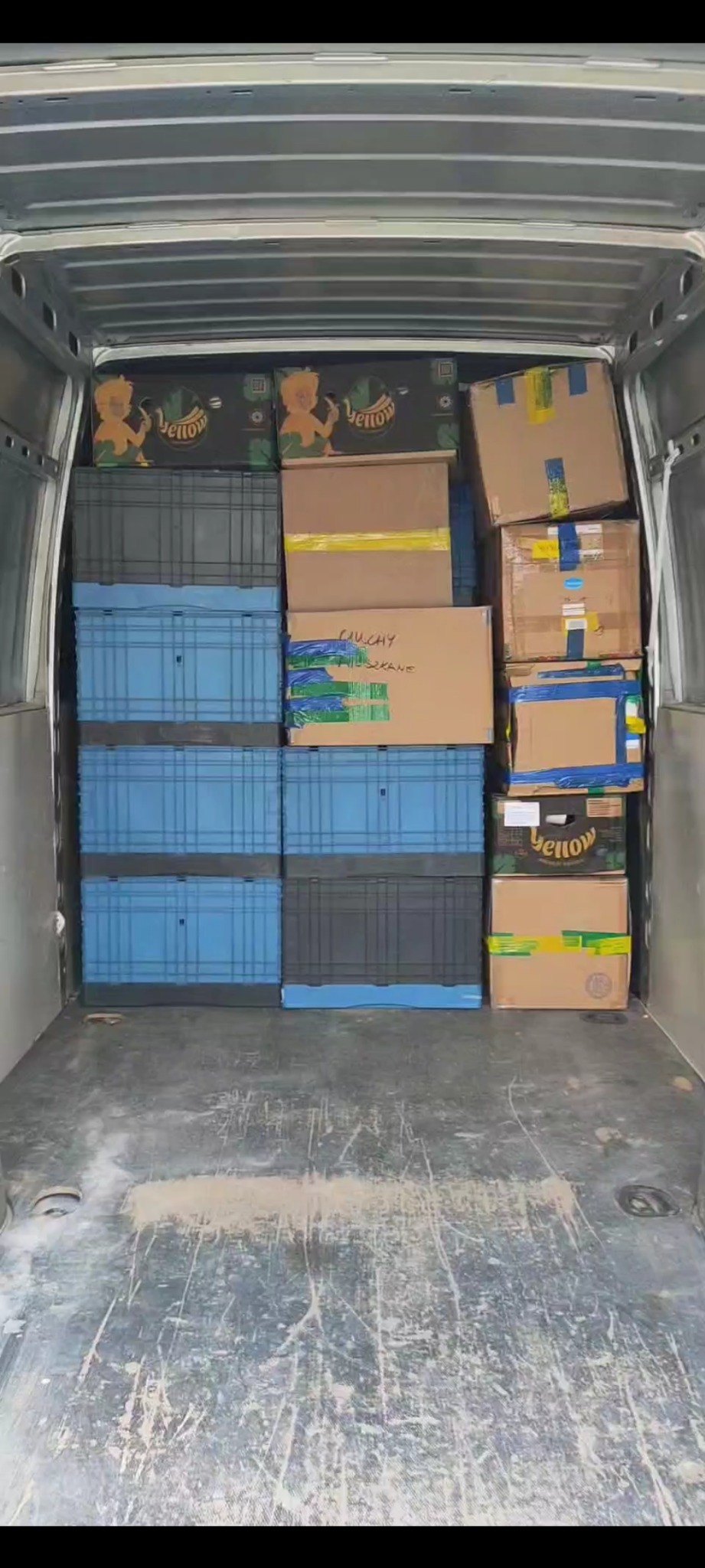 Wnętrze busa wypełnione kartonami i niebieskimi skrzynkami, gotowe do transportu. Widok z tyłu pojazdu, podłoga lekko zabrudzona.