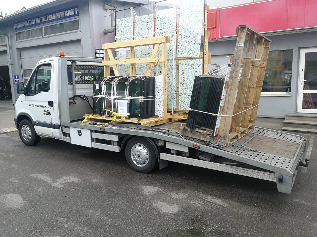 Laweta z ładunkiem szkła i drewnianych konstrukcji na platformie, zaparkowana przed budynkiem stacji kontroli pojazdów. Widoczny numer telefonu.