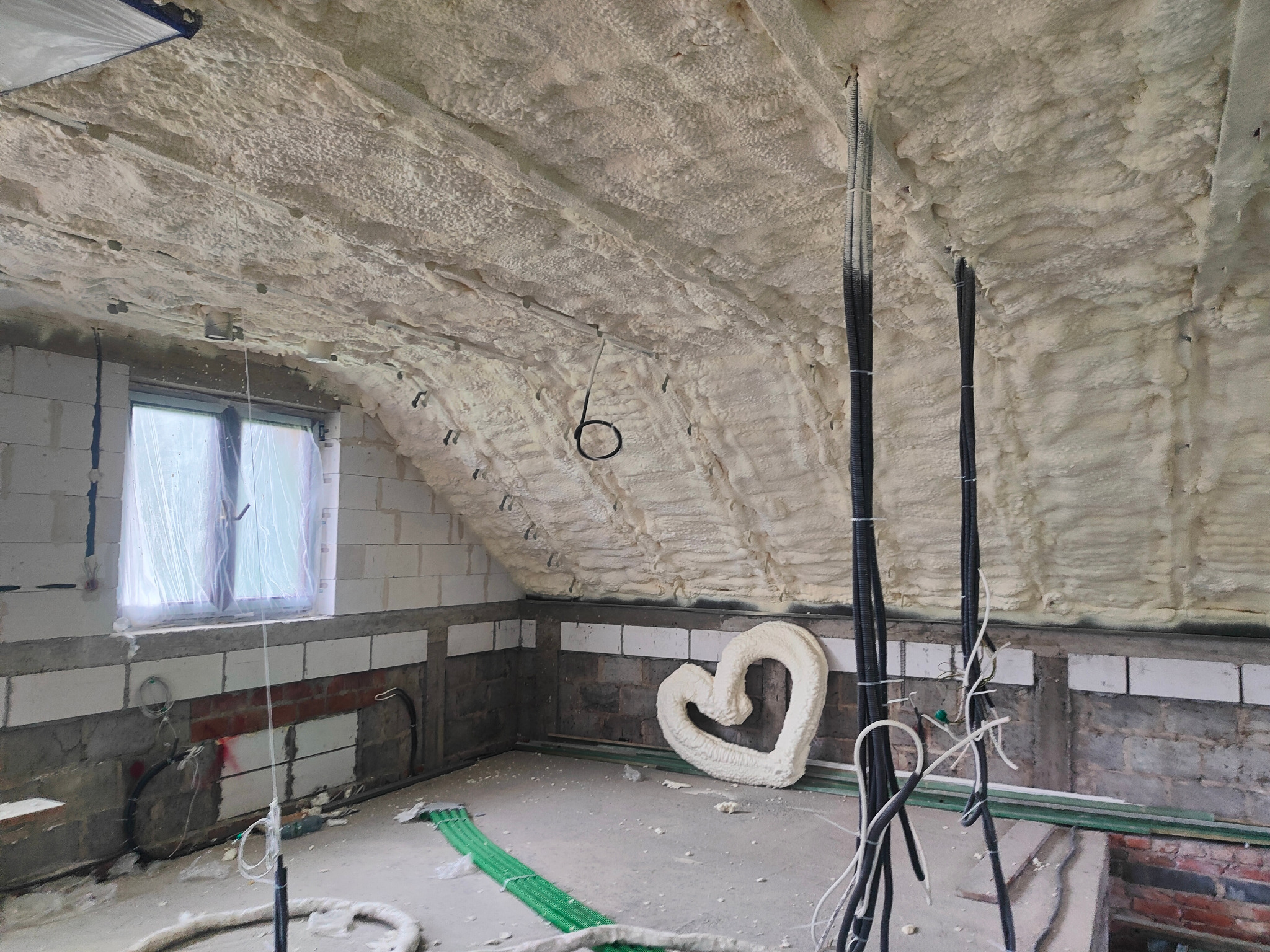Poddasze po ociepleniu pianką PUR, widoczne przewody elektryczne i kształt serca z pianki. Okno z folią w tle. Ściany z częściowo wykończoną cegłą.
