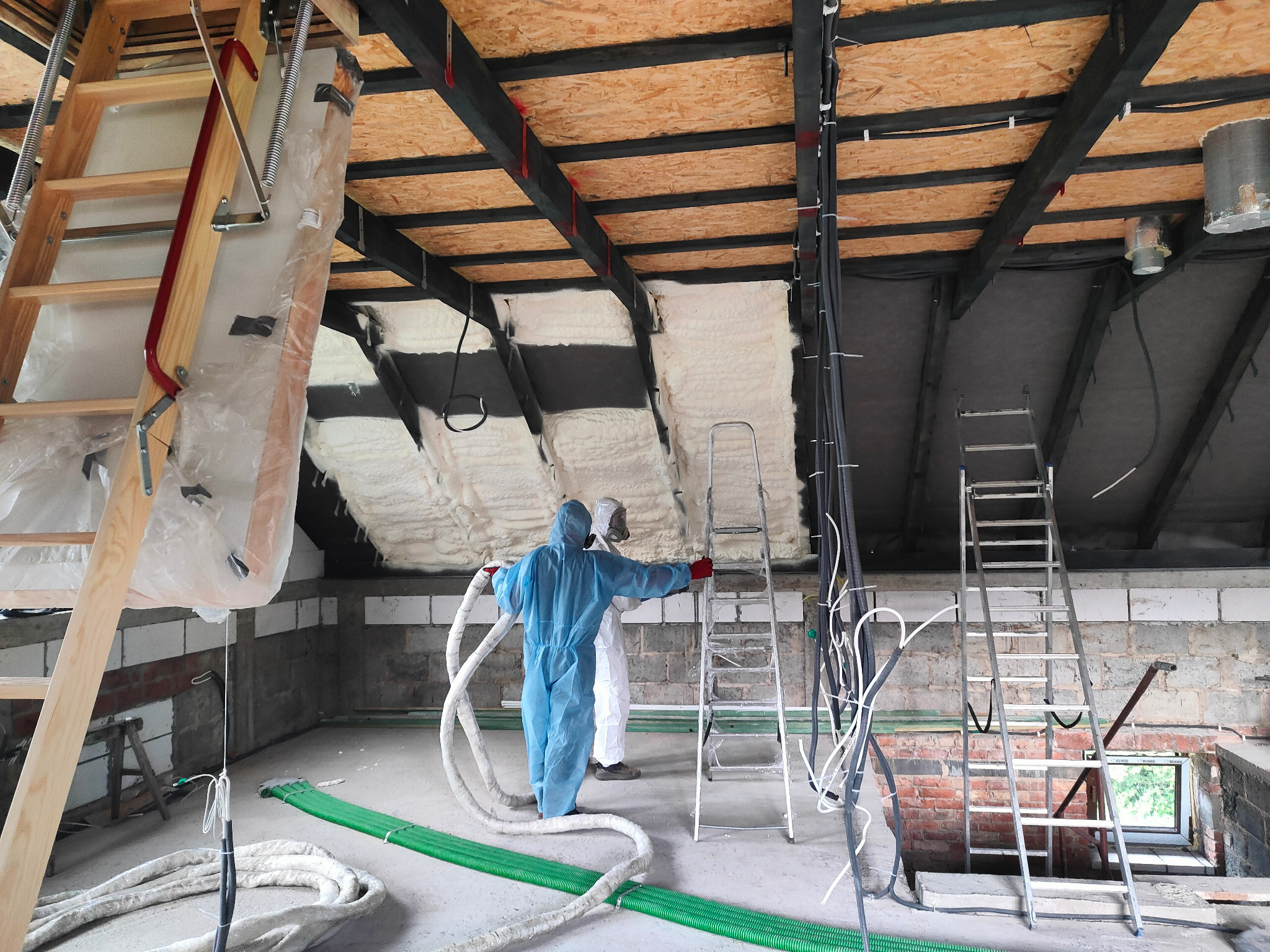 Ocieplanie poddasza pianką PUR w trakcie realizacji. Dwóch pracowników w kombinezonach aplikuje pianę na skośny dach. Widoczne drabiny, przewody i elementy konstrukcyjne.