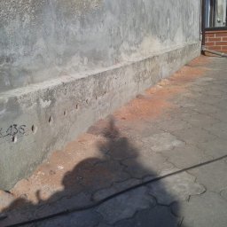 HYDROSYSTEM BESKID SPÓŁKA Z OGRANICZONĄ ODPOWIEDZIALNOŚCIĄ - Fundament budynku z nawierconymi otworami, częściowo odsłonięty, widoczny fragment elewacji i chodnika z kostki brukowej.