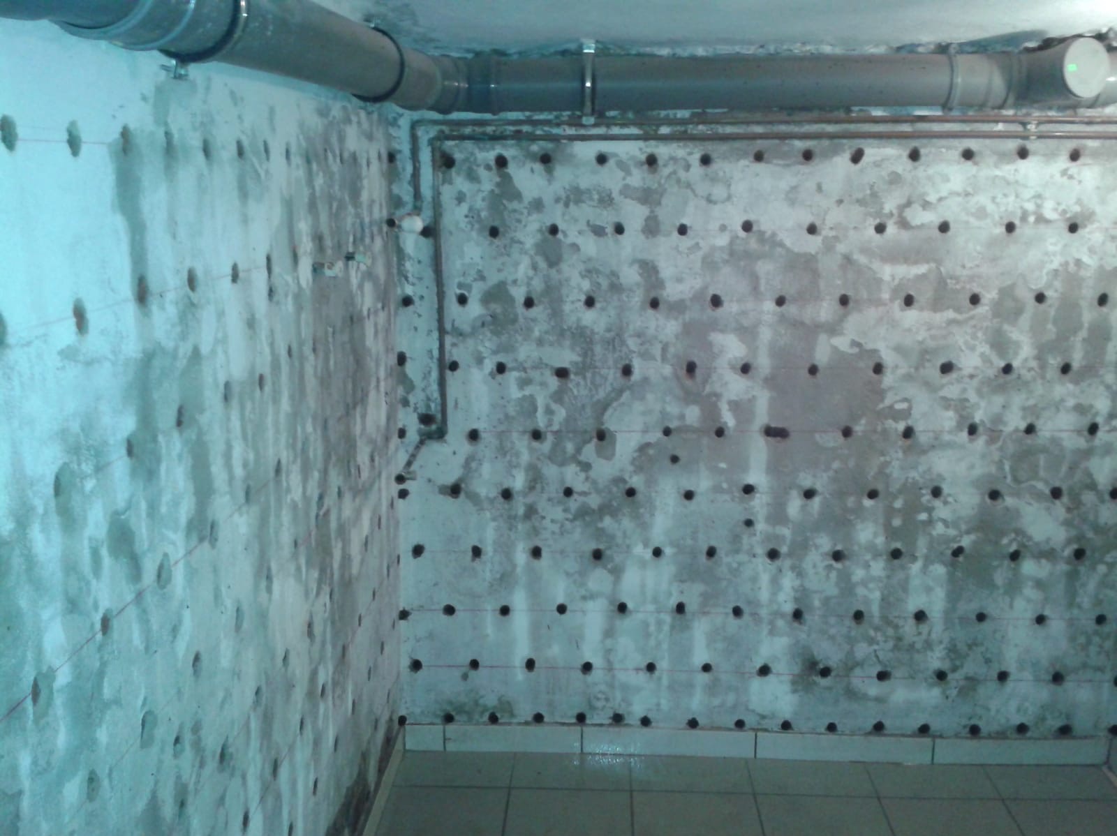 Wilgotna ściana piwnicy z regularnie rozmieszczonymi otworami, prawdopodobnie przygotowana do iniekcji osuszającej, widoczne rurki instalacyjne pod sufitem.