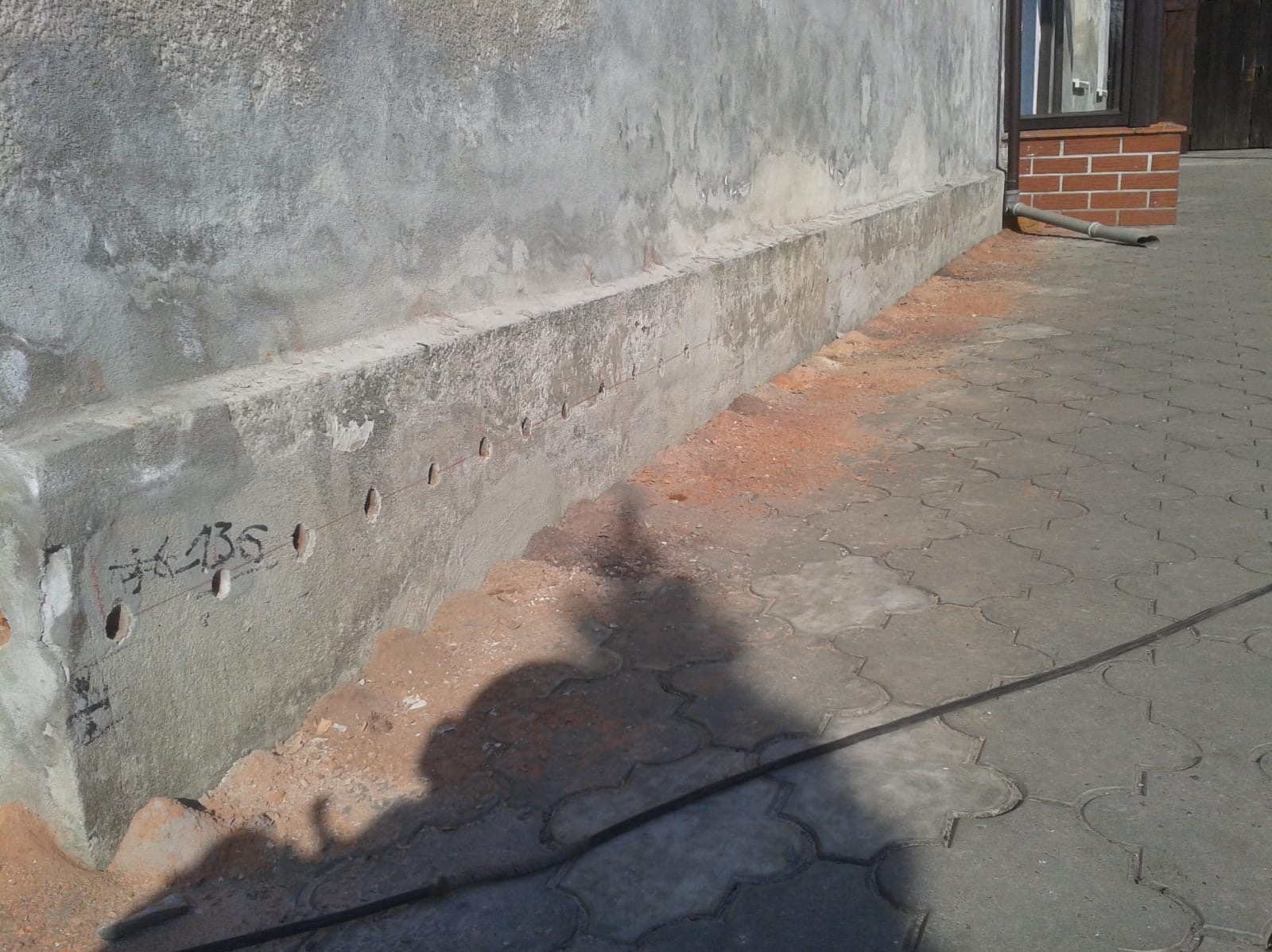 Fundament budynku z nawierconymi otworami, częściowo odsłonięty, widoczny fragment elewacji i chodnika z kostki brukowej.