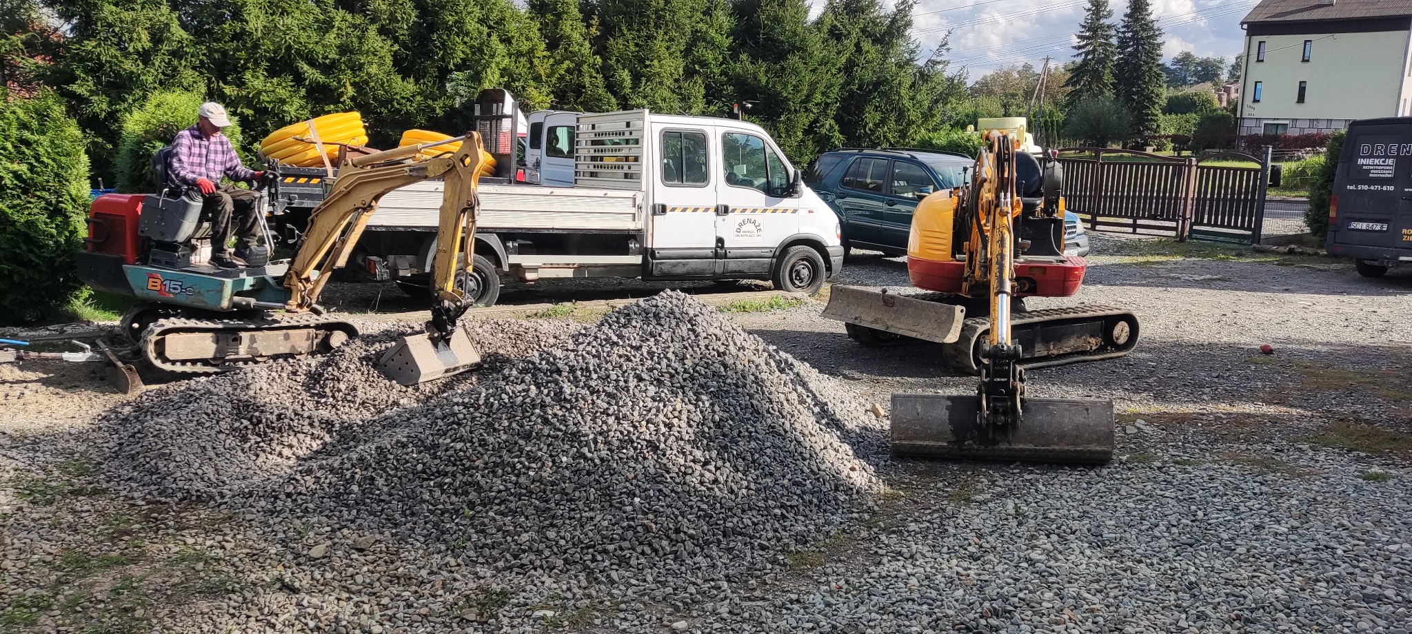 Dwa mini-koparki, jedna z operatorem w kraciastej koszuli, pracują na żwirowym terenie. Na pierwszym planie spory stos żwiru, w tle biały samochód dostawczy i zieleń.