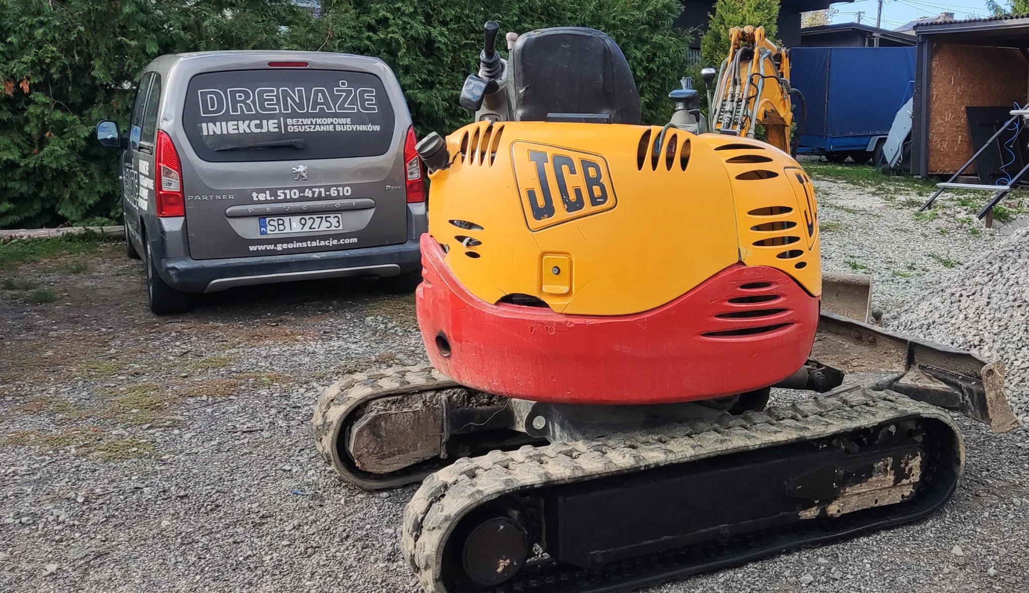 Żółto-czerwona mini koparka JCB na żwirowym podłożu, z tyłu szary samochód dostawczy z reklamą drenażu, iniekcji i osuszania budynków oraz sterta żwiru.