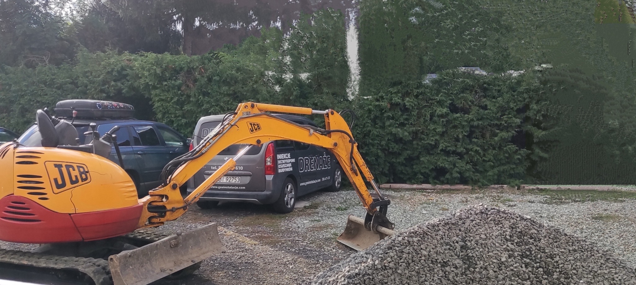 Żółto-czerwona mini koparka JCB na żwirowym terenie, w tle samochody osobowe i bus z napisem 'Drenaże' oraz danymi kontaktowymi firmy.