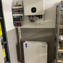 AM GROUP SOLAR MARCIN SERKOWSKI - Biały magazyn energii firmy Hinen z podłączonym inwerterem, zamontowany w rogu pomieszczenia z szarą, ziarnistą ścianą, obok metalowego regału z słoikami i butelkami.