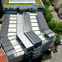 Widok z góry na budynek z panelami słonecznymi na dachu, z zielonym dachem na sąsiednim budynku, otoczony ulicami i drzewami.
