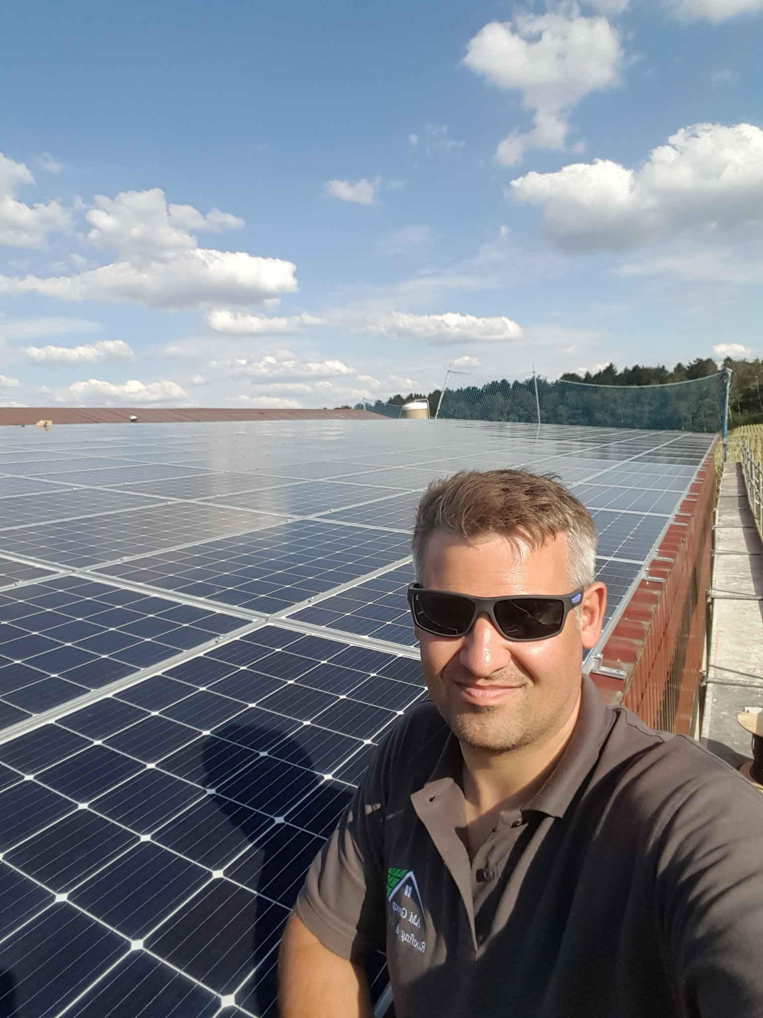 Instalator w okularach przeciwsłonecznych, pozujący na tle dużej instalacji fotowoltaicznej na płaskim dachu z widoczną siatką zabezpieczającą i błękitnym niebem z chmurami.