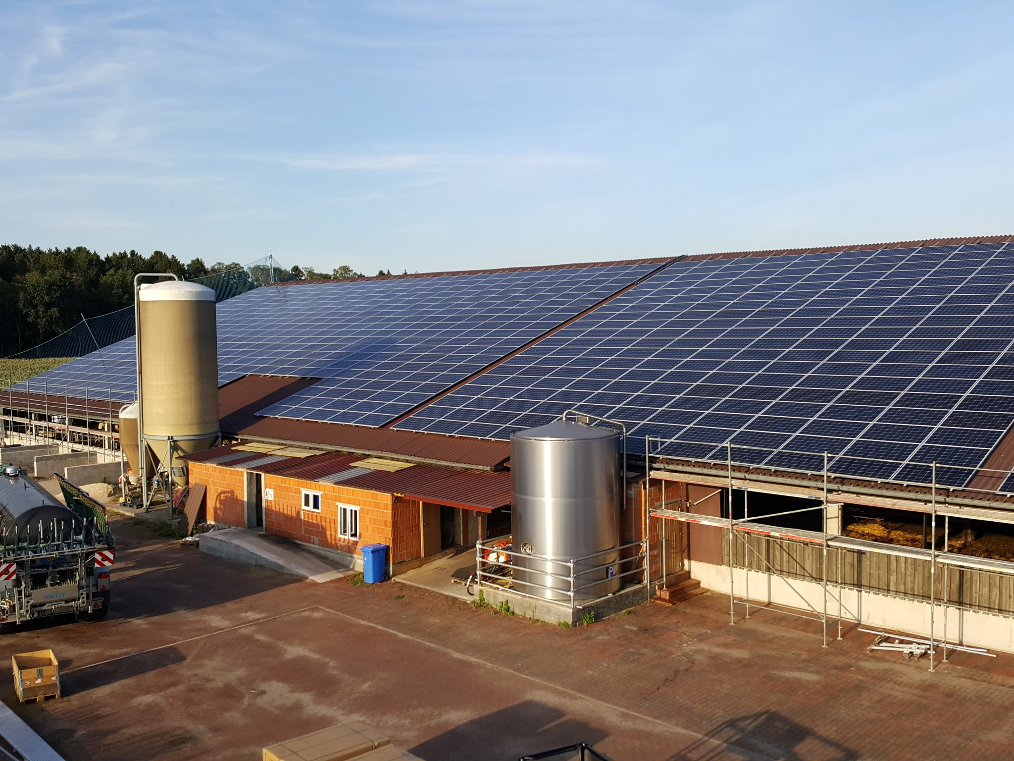 Instalacja paneli słonecznych na dachu budynku gospodarczego z dwoma silosami i zaparkowanym pojazdem rolniczym, widok z góry.