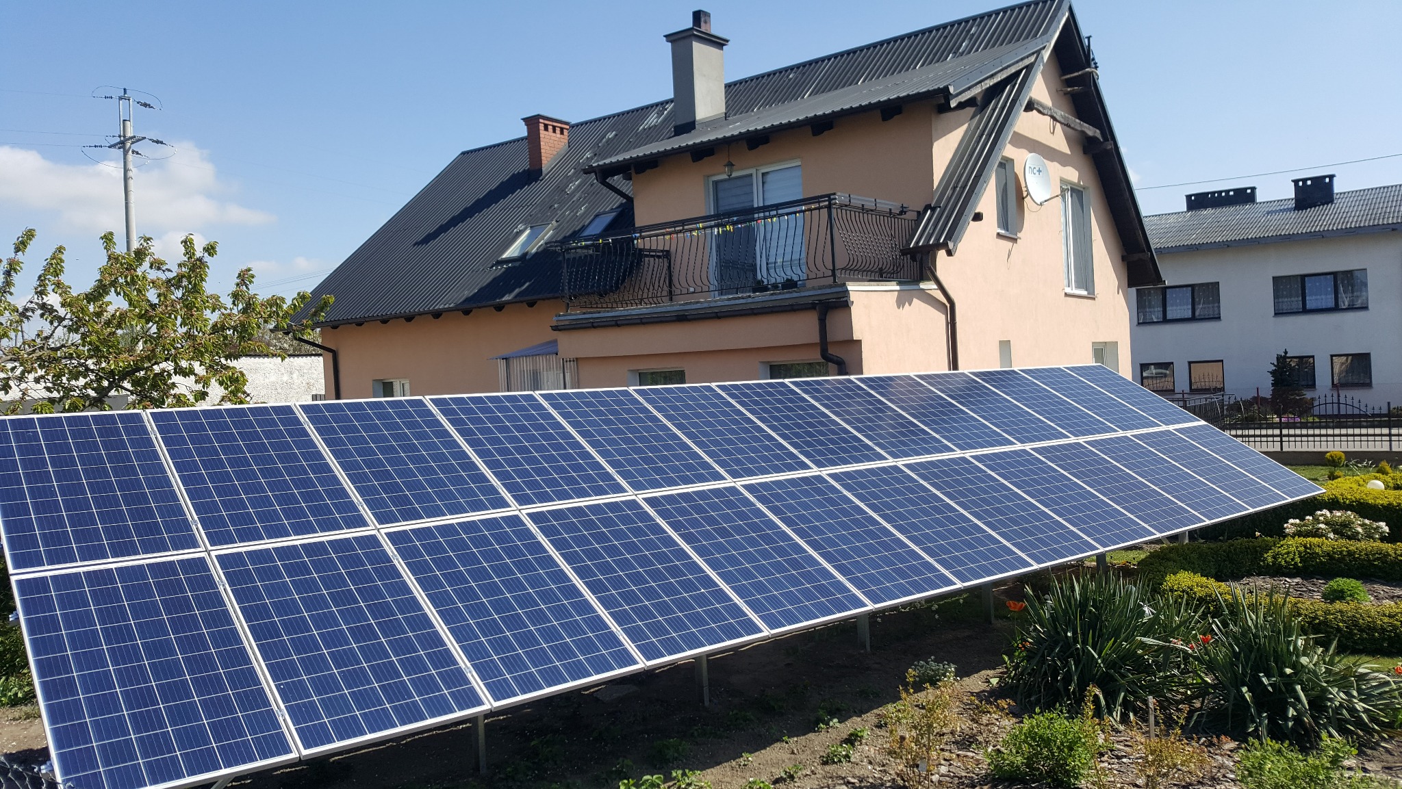 Gruntowa instalacja paneli słonecznych na tle domu z balkonem i ogrodem w słoneczny dzień.