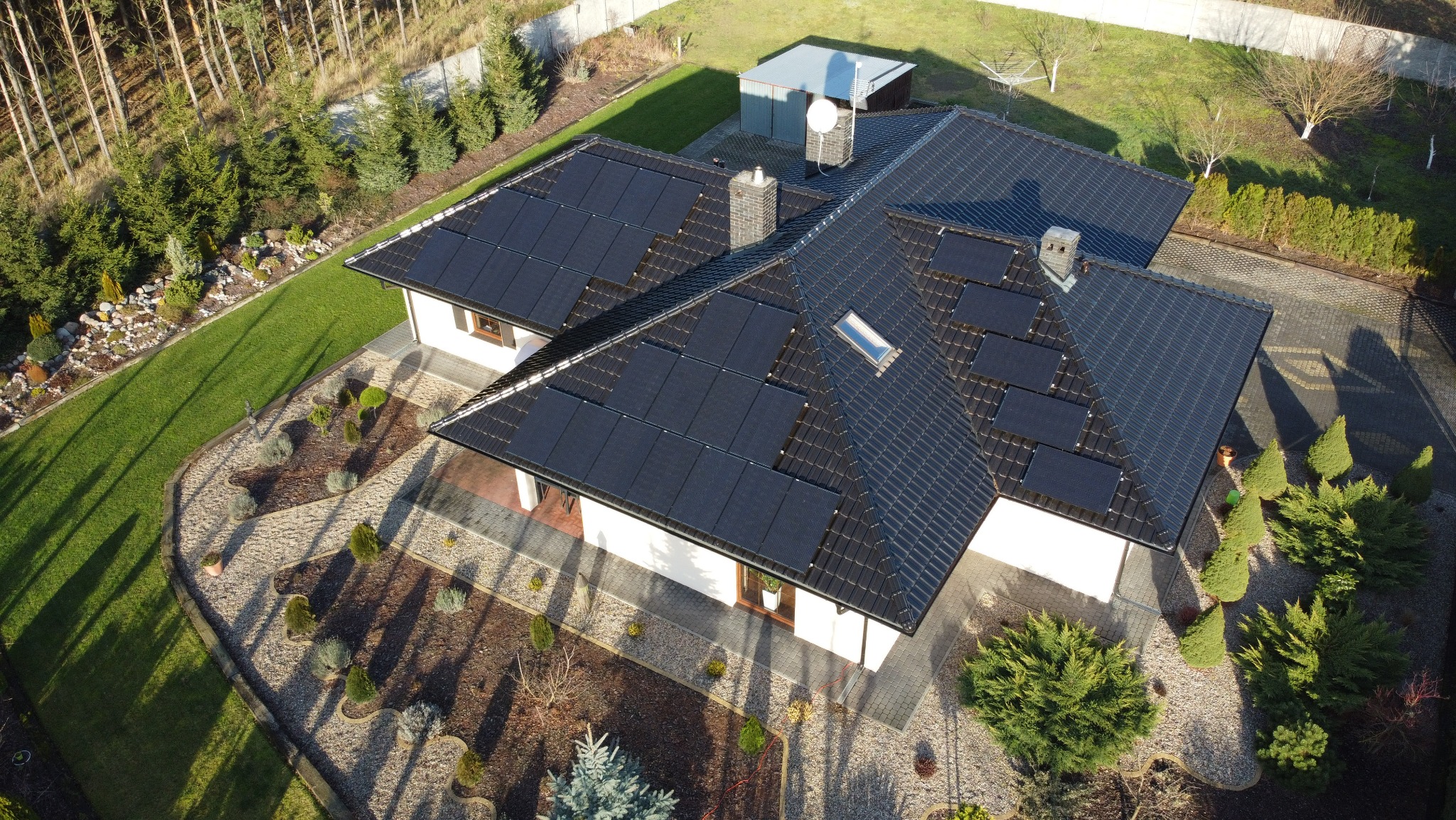 Widok z lotu ptaka na dom jednorodzinny w Ostróżkach z zainstalowanymi panelami słonecznymi na dachu, otoczony zadbanym ogrodem z licznymi krzewami i drzewami iglastymi. Na dachu widoczny komin...