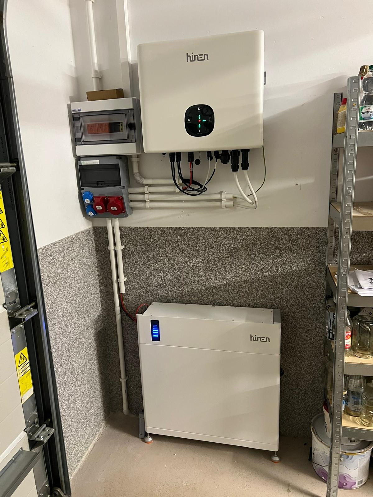 Biały magazyn energii firmy Hinen z podłączonym inwerterem, zamontowany w rogu pomieszczenia z szarą, ziarnistą ścianą, obok metalowego regału z słoikami i butelkami.