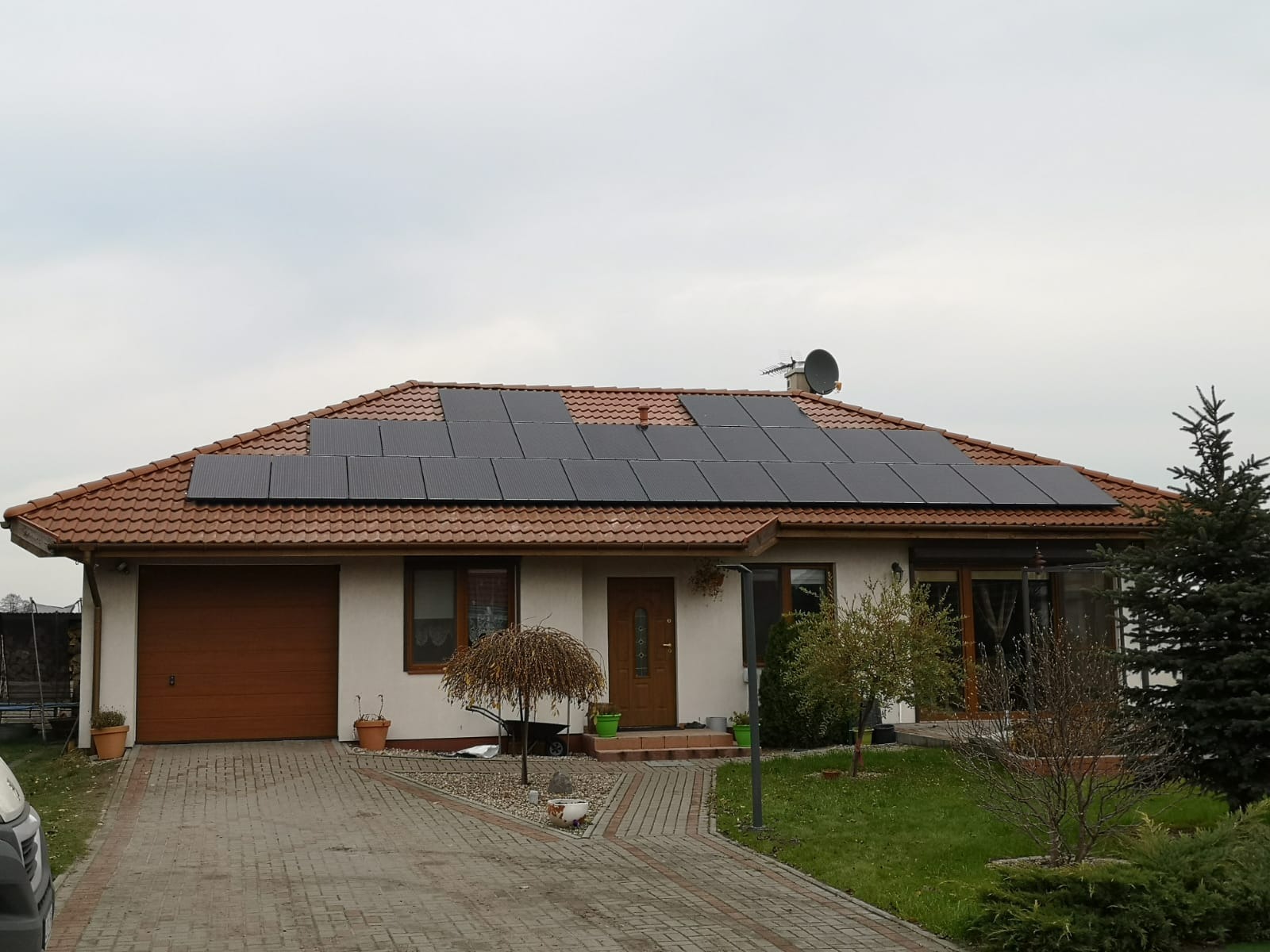 Dom jednorodzinny z brązową dachówką i panelami słonecznymi na dachu, widok z chodnika z kostki brukowej w pochmurny dzień.