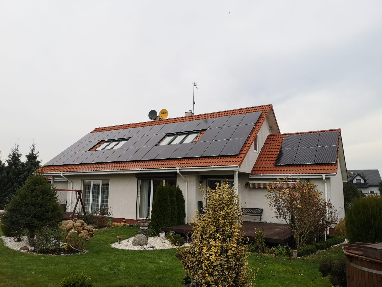 Dom jednorodzinny z panelami fotowoltaicznymi na dachu z czerwonej dachówki, widoczne okna dachowe i dwie anteny satelitarne, ogród z elementami dekoracyjnymi.