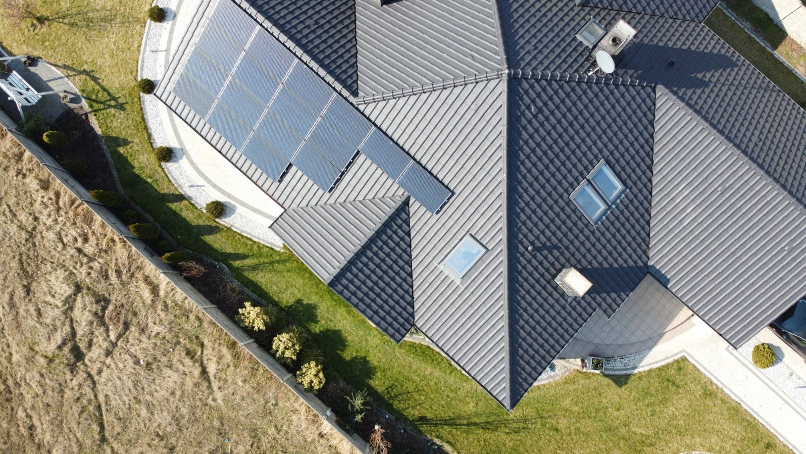 Widok z góry na dom z szarym dachem i panelami słonecznymi, otoczony zielonym trawnikiem i ozdobnymi krzewami, z widocznym fragmentem ogrodzenia i samochodu.