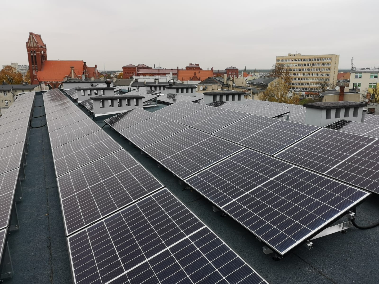 Rozległa instalacja fotowoltaiczna na dachu budynku miejskiego, widok z góry, w tle czerwona wieża kościoła i zabudowa miejska w pochmurny dzień.