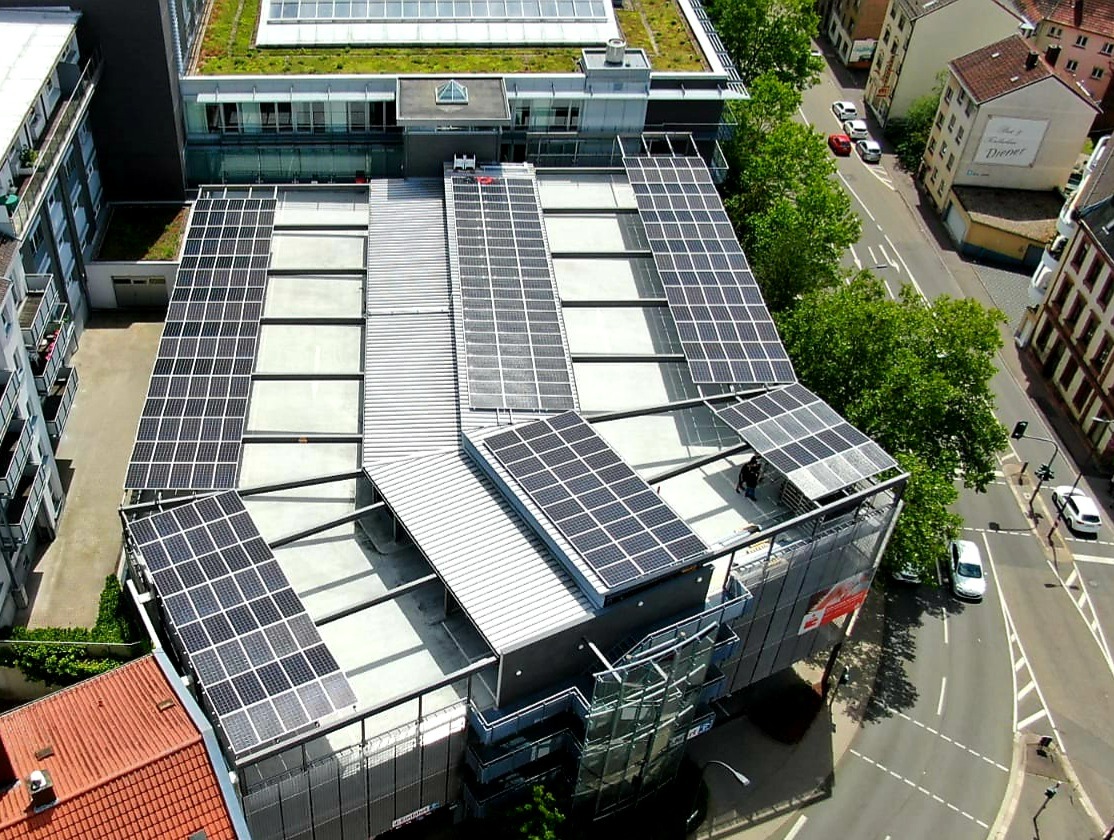Widok z góry na budynek z panelami słonecznymi na dachu, z zielonym dachem na sąsiednim budynku, otoczony ulicami i drzewami.