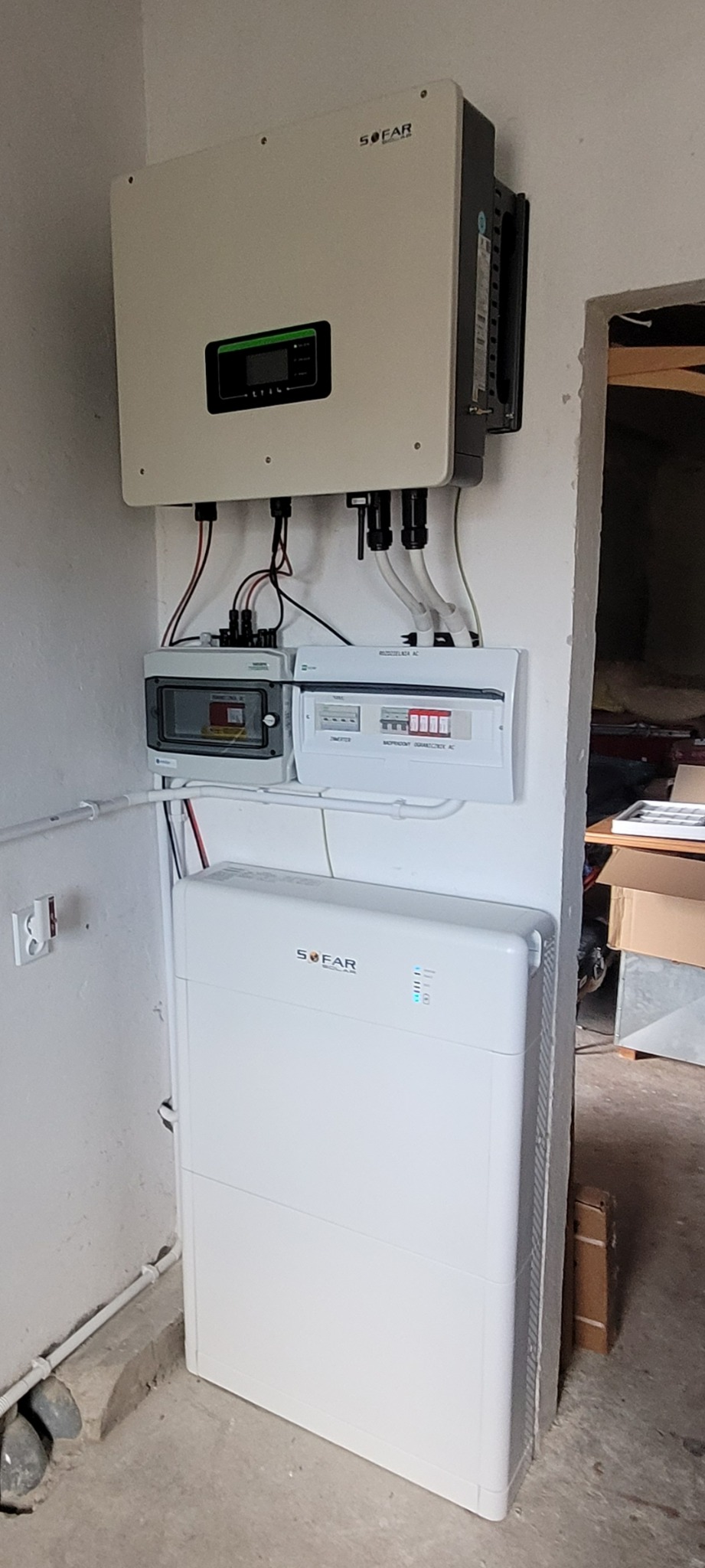 Biały magazyn energii Sofar połączony z inwerterem i rozdzielnicami na ścianie w surowym pomieszczeniu.
