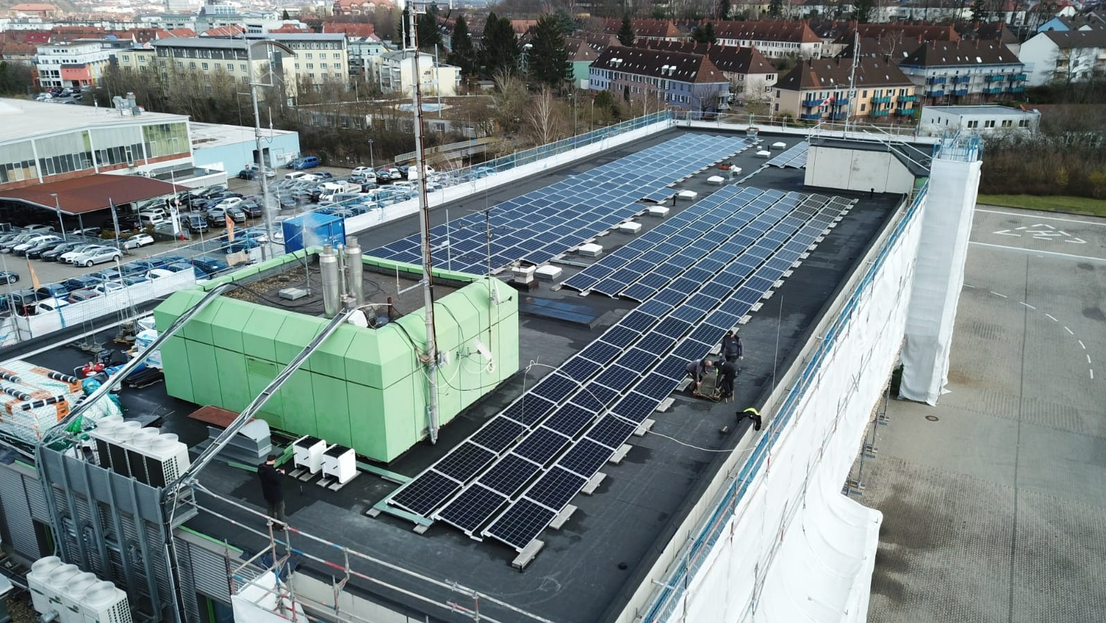 Instalacja paneli słonecznych na dachu budynku z zieloną konstrukcją, widok z góry na miasto i parking.