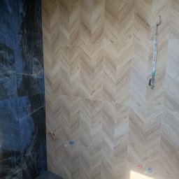 Michał Gwizdek - Ściana łazienki wyłożona płytkami imitującymi drewno w jodełkę, kontrastująca z ciemną, marmurkową płytką po lewej stronie. Widoczne wystające rury instalacyjne i smycz z kluczami wisząca na haczyku.