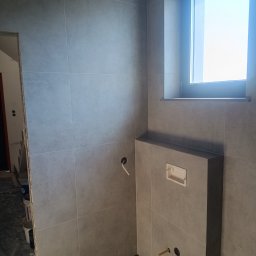Michał Gwizdek - Szare płytki ceramiczne na ścianie w łazience, z widocznym oknem i przygotowanym miejscem na stelaż podtynkowy WC z wystającymi rurami instalacyjnymi.