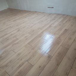 Michał Gwizdek - Ułożone panele podłogowe o wzorze jasnego drewna w nowym pomieszczeniu, widoczne gniazdka elektryczne w ścianie, ślady po szpachlowaniu na ścianach.