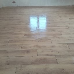 Michał Gwizdek - Ułożona podłoga z paneli imitujących drewno, z widocznym odbiciem światła okiennego, świeżo po montażu w surowym wnętrzu.