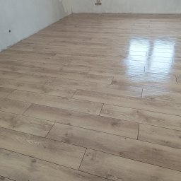 Michał Gwizdek - Jasne panele podłogowe z wyraźną strukturą drewna, ułożone w nowym pomieszczeniu z surowymi ścianami, odbijające światło okna.