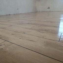 Michał Gwizdek - Świeżo ułożone, błyszczące panele podłogowe o wzorze drewna w jasnym odcieniu, z widocznymi szczelinami dylatacyjnymi i surową, niepomalowaną ścianą w tle.