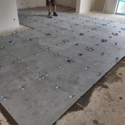 Michał Gwizdek - Układanie szarych płytek podłogowych z użyciem systemu poziomowania, widoczne krzyżyki dystansowe i kliny, fragment podłogi niepokryty płytkami.