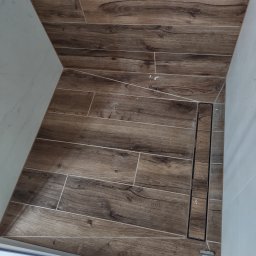 Michał Gwizdek - Wykończona kabina prysznicowa z brązowymi płytkami imitującymi drewno i liniowym odpływem, widoczne białe ściany i fragment kabiny.